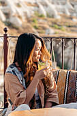 Ethnische Frau mit langen gewellten Haaren sitzt auf einer Terrasse und nippt an einem erfrischenden Cocktail aus einem Glas mit Strohhalm, während sie die Landschaft auf der Terrasse in Kappadokien, Türkei, bewundert