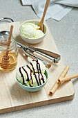 Von oben leckere Eiskugeln in einer Schüssel mit Schokoladensoße, serviert auf einem hölzernen Schneidebrett mit einem Glas Honig und Waffelröllchen in der Küche