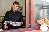 Positive adult male in black outfit holding plate with tasty salad while standing in restaurant and looking at camera