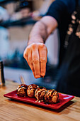 Gesichtsloser Koch gießt mit Gewürzen Fleischröllchen mit Soße auf einen stilvollen Teller in einer Ramen-Bar
