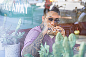 Through window view of young dreamy male in sunglasses enjoying lunch in disposable wrapper while looking away