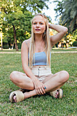 Ruhige Frau in Sommerkleidung sitzt auf einer grünen Wiese im Garten und schaut in die Kamera