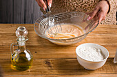 Crop anonymous female whisking eggs while preparing batter for crepes at wooden table with oil and flour in light kitchen
