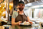 Female worker in protective mask looking at camera and serving dessert while standing at counter during work in modern cafe