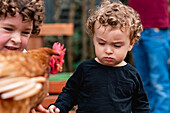 Cute little curly haired boy showing hen to crop brother in garden in agricultural farm