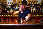 Young Asian bartender pouring strawberry juice in the shaker while preparing a cocktail in the bar