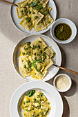 Draufsicht auf appetitlich gekochte Ravioli mit grüner Soße und Kräutern auf weißen Tellern mit Gabeln auf dem Tisch