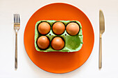 Draufsicht auf eine Kartonschachtel mit Eiern auf einem orangefarbenen Keramikteller auf einem weißen Tisch mit Messer und Gabel