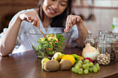 Happy asian woman in casual clothes stirring salad with assorted veggies and fruits with wooden spoon in bowl while cooking in kitchen on sunny day