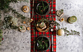 Top view of Christmas table setting with wreath on the plate, decorative wooden ornaments and red checkered tablecloth with yellow lights on the background