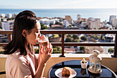 Glückliche Asiatin mit einem Glas Heißgetränk in der Hand, die in die Kamera schaut, während sie beim Frühstück auf dem Balkon am Tisch sitzt