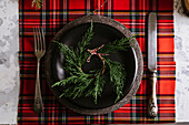 From above Christmas table setting with wreath on ceramic plate with cutlery on red checkered tablecloth on the background