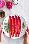 Top view of crop unrecognizable person showing gravlax with mixed peppercorns and fresh dill sprigs on plate on light background