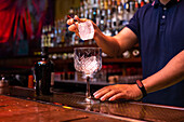 Unrecognizable bartender putting a big ice cube into the glass while preparing a gin tonic cocktail in the bar