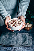 Hand of crop unrecognizable person holding a bowl with raw chickpeas on table with towel and green plants in light kitchen