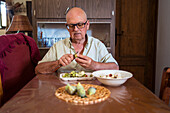 Älterer Mann mit Brille und Messer schält eine grüne Feige an einem Tisch mit einem Einwegtablett in einem Hausraum