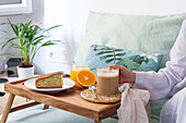 Unrecognizable crop female sitting on bed with cup of coffee and tray served with sponge cake and glass of juice while having breakfast at home