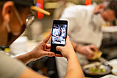 Cropped waitress in uniform and protective mask taking photo on smartphone of chef and cooks preparing food in open kitchen during coronavirus outbreak
