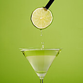 Eine nicht erkennbare Person hält eine frische Limettenscheibe über einem Glas mit einem Daiquiri-Cocktail vor grünem Hintergrund