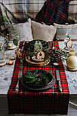 Weihnachtlich gedeckter Tisch mit Kranz auf dem Teller, dekorativem Holzschmuck und rot kariertem Tischtuch mit gelben Lichtern im Hintergrund