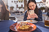 Focused Asian female taking picture of tasty shrimp dish on smartphone while sitting at table with friends during lunch in outdoor cafeteria