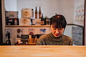 Zufriedene asiatische Frau in legerem Pullover, die an einem Holztisch in einer Ramen-Bar sitzt und nach unten schaut