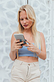 Ruhige Frau mit blondem Haar und in Sommerkleidung steht in der Stadt und surft auf ihrem Handy