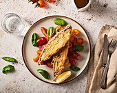 Top view of appetizing dish with whole pan fried mackerel placed on cut cherry tomatoes decorated with fresh basil leaves and served on table with water glass and cutlery