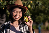 Delighted ethnic female farmer standing with ripe bitten pear in summer garden in countryside and looking at camera