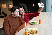 Fröhliches, junges, gemischtrassiges Paar sitzt am Tresen mit gesundem vegetarischem Salat und Getränken und macht ein Selfie mit dem Smartphone beim gemeinsamen Frühstück in einem modernen Restaurant