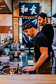 Side view of male chef in black uniform and bandana cooking Asian dish called ramen in modern cafe