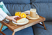 High angle of crop faceless female taking piece of homemade sponge cake from tray with cup of coffee and glass of orange juice during breakfast at home