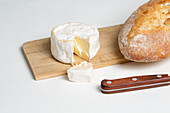 Leckerer Camembert-Käse auf einem hölzernen Schneidebrett neben Brot und Messer auf weißem Hintergrund