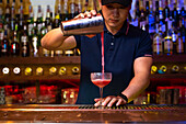 Young Asian bartender working in the bar with his shaker and pouring a cocktail in the glass