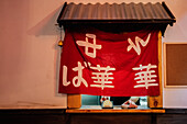 Bildausschnitt Person, die ein asiatisches Gericht mit Soße auf ein Holzbrett mit rotem Tuch am Fenster eines Cafés stellt