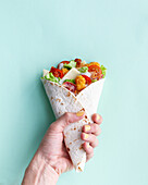 Top view of cropped unrecognizable female holding delicious chicken wrap with fresh lettuce and ripe tomatoes in tortilla placed on light background in studio