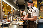 Seitenansicht eines männlichen Kochs mit Schutzmaske und Schürze, der einen Gasbrenner benutzt, während er Knochenmark brät, während er in einem Café arbeitet