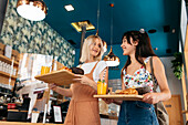 From below of young content girlfriends looking at each other while standing with delicious food and refreshing drinks in cafeteria