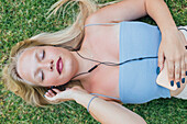 Top view of dreamy female enjoying songs in earphones while lying with closed eyes on grass in park in summer