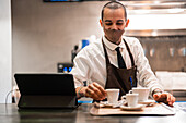 Positiver Kellner in Uniform und Maske, der Tassen mit aromatischem Brühkaffee auf ein Tablett am Tresen mit Tablet-Tastatur-Dock in einem Restaurant während einer Coronavirus-Pandemie stellt