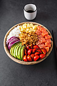 Draufsicht auf eine appetitliche bunte Poke-Schale mit frischem Lachs und Reis, serviert mit reifen Kirschtomaten, Avocadoscheiben, Zwiebeln und Mango auf einer Betonfläche mit Sojasauce