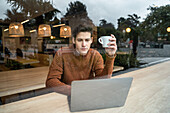Junger männlicher Student sitzt mit Laptop und einer Tasse Kaffee am Tisch und verbringt seine Zeit in einer modernen Cafeteria durch das Fenster