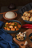 Mala-Tofu, chinesisches veganes Gericht, begleitet von einer Schüssel Reis, Blumenkohl, Sojasauce und einer japanischen Teekanne auf einem mit Stoffen dekorierten Holztisch