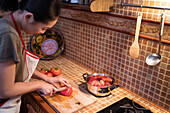 From above side view of ethnic female in apron cutting ripe tomatoes on chopping board while cooking lunch in kitchen at home