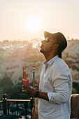 Side view of trendy African American male in sunglasses with bottle of beer puffing on balcony at sunset in Cappadocia, Turkey