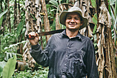 Positive Hispanic man in shirt and bucket hat touching shoulder with machete and looking at camera with smile during work on ecological banana farm in Costa Rica