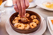 From above of crop unrecognizable woman dipping bread piece into sauce of appetizing traditional gambas al ajillo dish served in bowl in restaurant