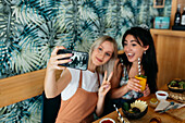 Young cheerful best female friends taking self portrait on cellphone while showing victory gesture during brunch in cafe