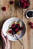 Top view of anonymous person taking bowl of delectable crepes with blueberry jam and raspberries from timber table during breakfast