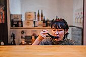Young Asian woman in sweater eating ramen with spoon at wooden counter in cafe